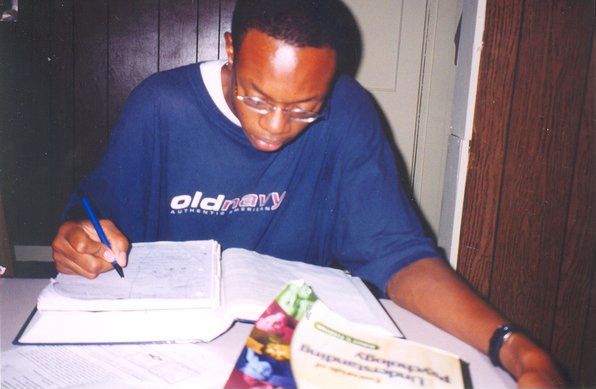 Big forehead guy (possibly young Odinaka?) looking at book while at desk with pen and notebook in hand